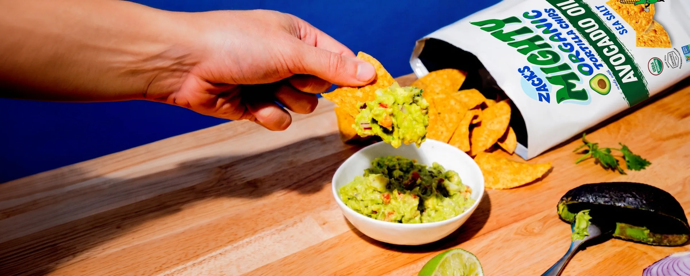 A hand lifting a chip loaded with guacamole beside an open bag of Zack's Mighty chips